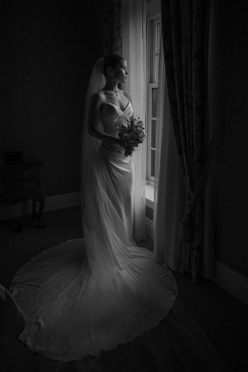 Elegant bride in a flowing white gown and veil holding a bouquet, standing by a window in a black and white portrait.