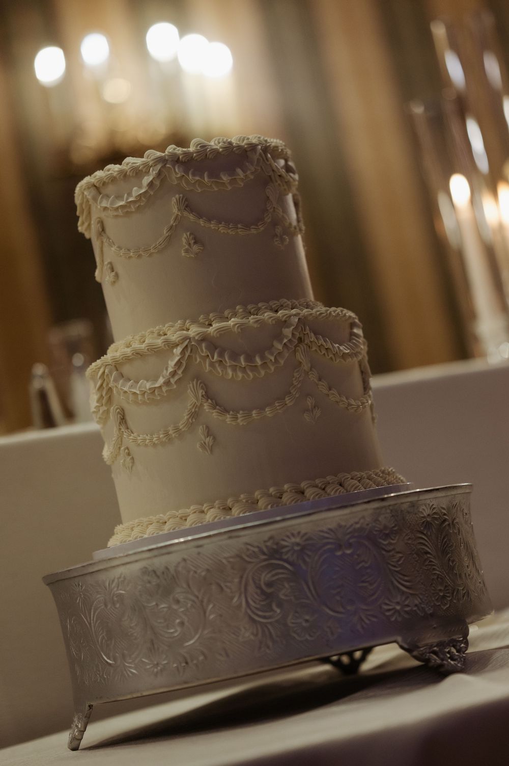 Elegant three-tier white wedding cake with pearl and swag decorations on a silver ornate stand.