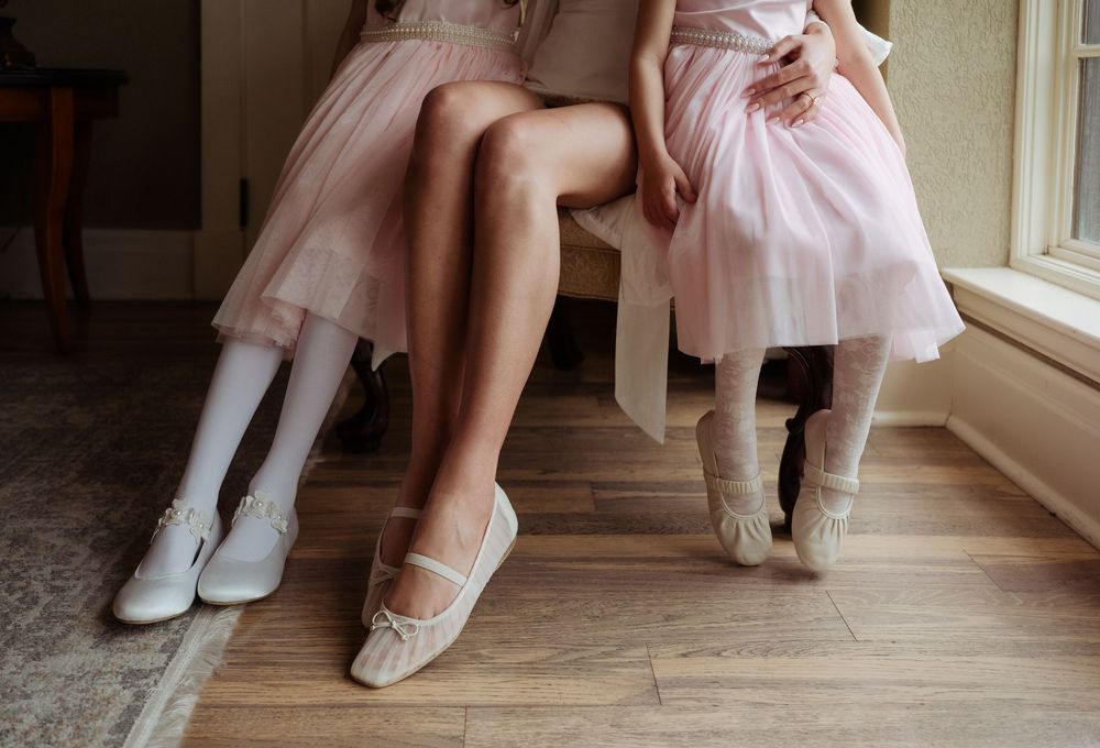 Two females in pink dresses sit together, one wearing white tights and flats, the other in ballet slippers, on a wooden floor.