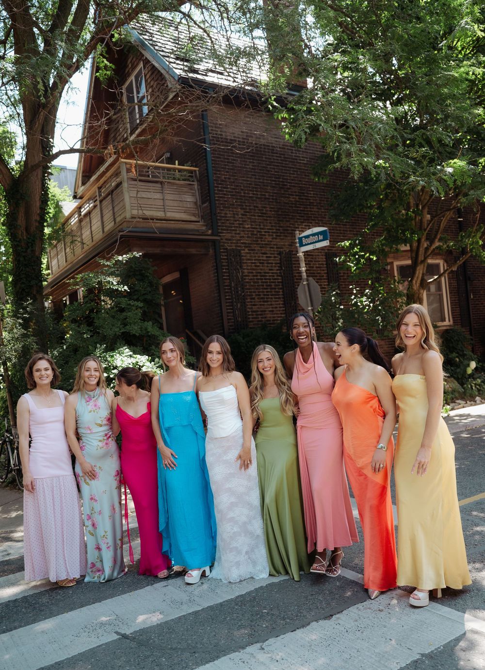 A bride in white stands with eight bridesmaids in colorful rainbow dresses on a street outside a brick house.
