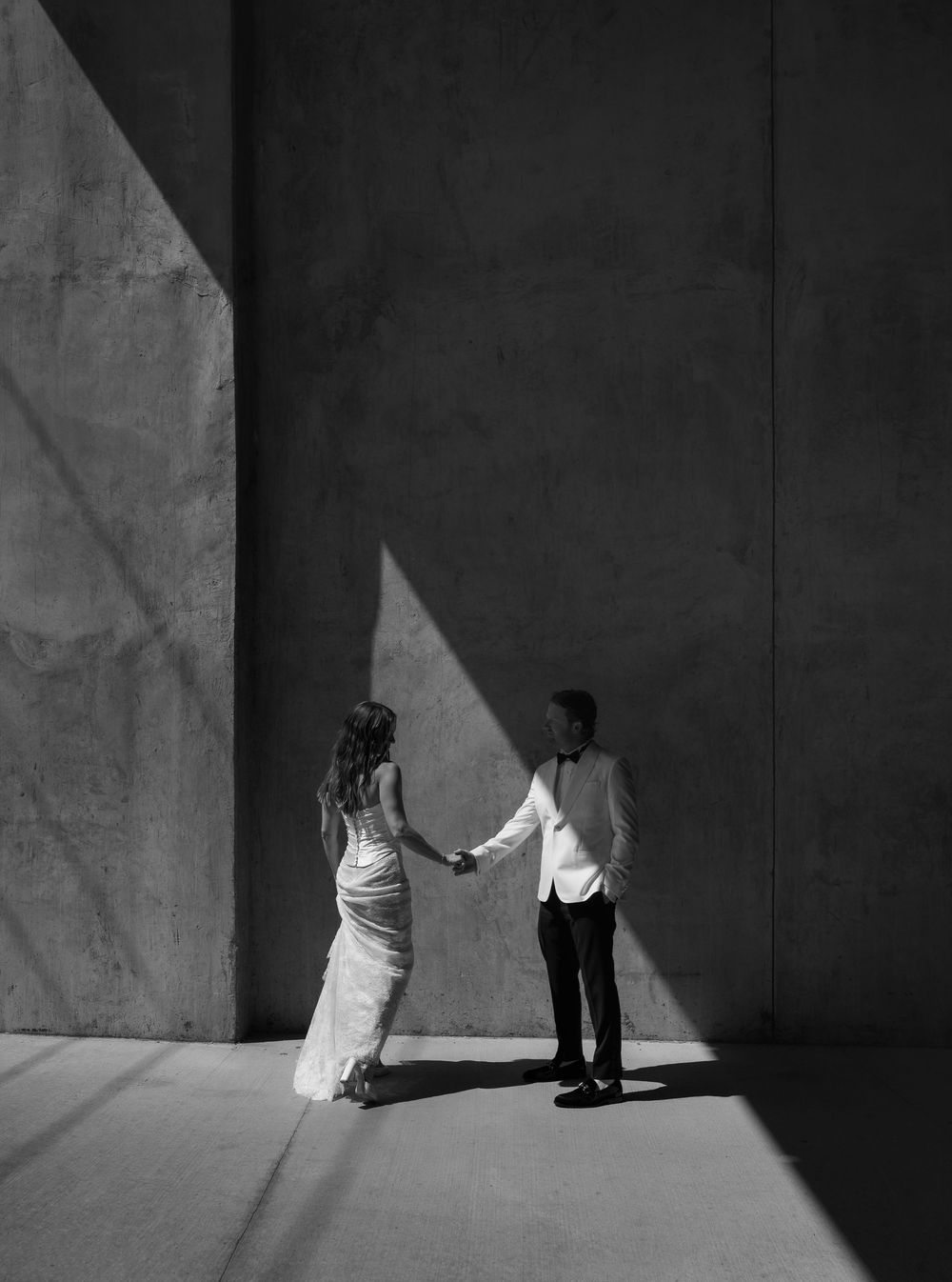 Black and white photo of a couple holding hands in dramatic light against a concrete wall.