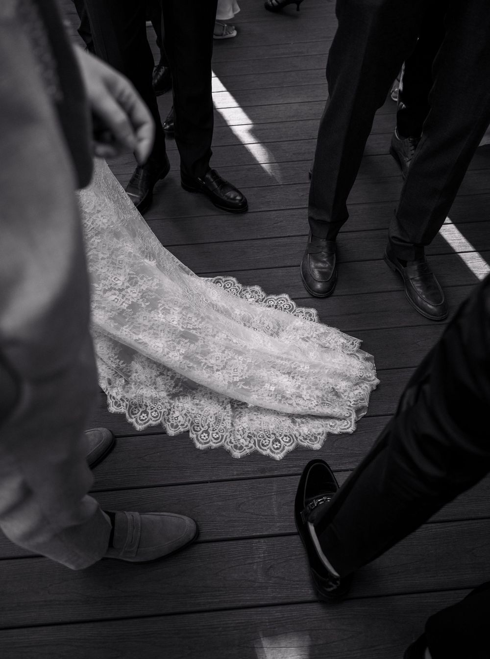 Black and white photo of a bride's lace wedding dress train on wooden floor surrounded by guests in formal shoes.