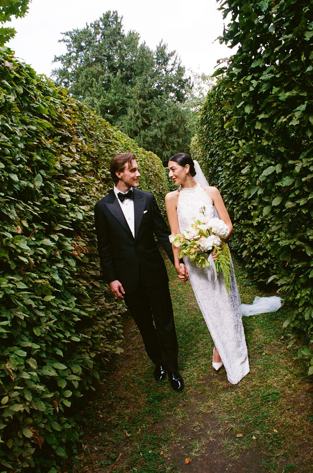A couple in formal wedding attire walks through a lush garden path surrounded by green hedges and foliage.