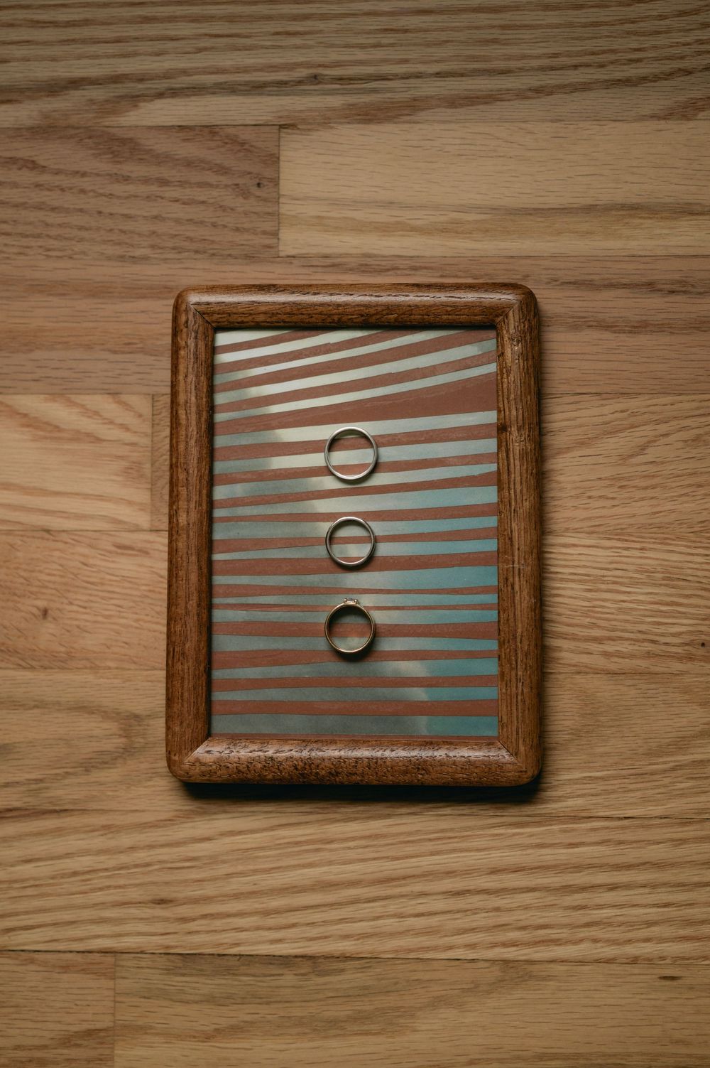 Wedding rings displayed on a blue striped background in a wooden frame create an artistic composition.
