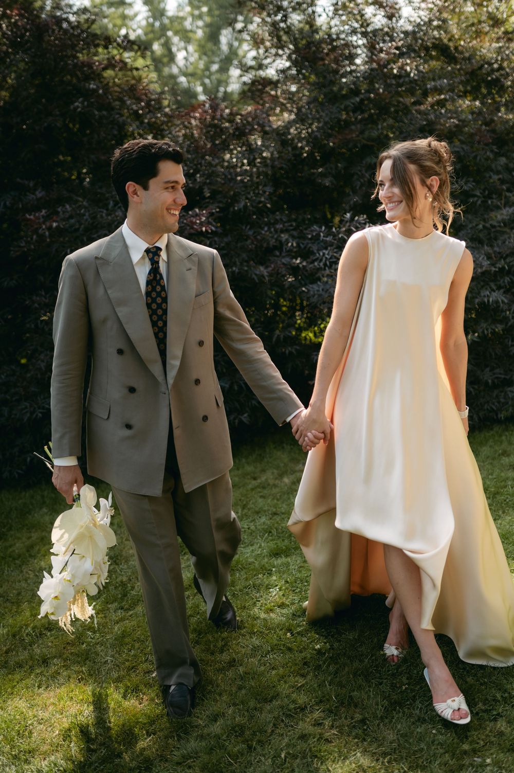 A couple in wedding attire walks together on grass during a sunny outdoor photo session.