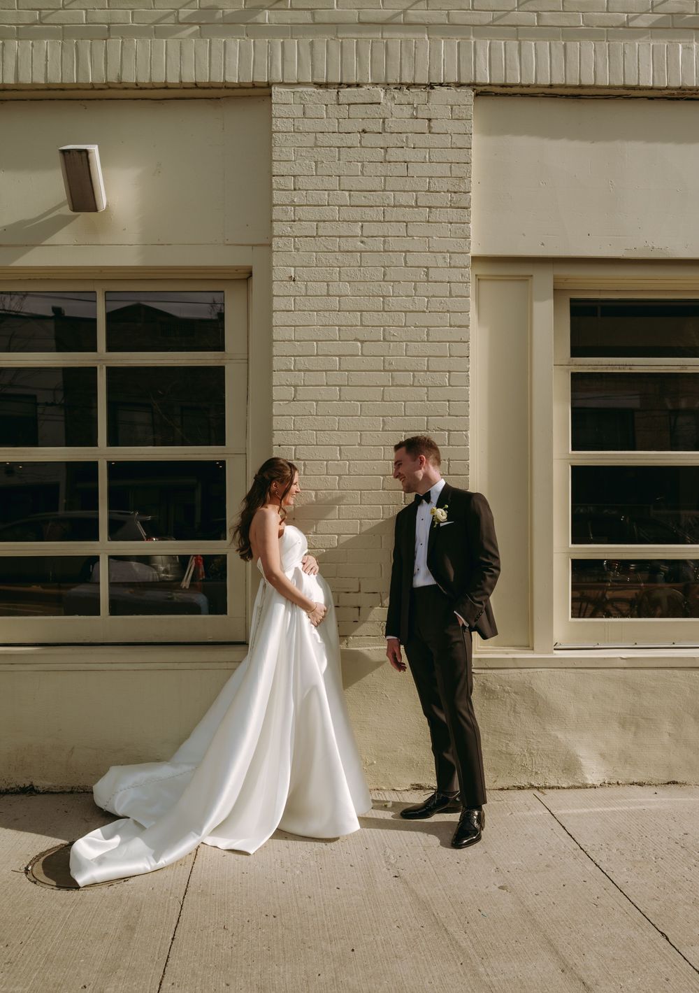 Bride in white gown and groom in black tuxedo share a romantic moment outside a brick building on their wedding day.