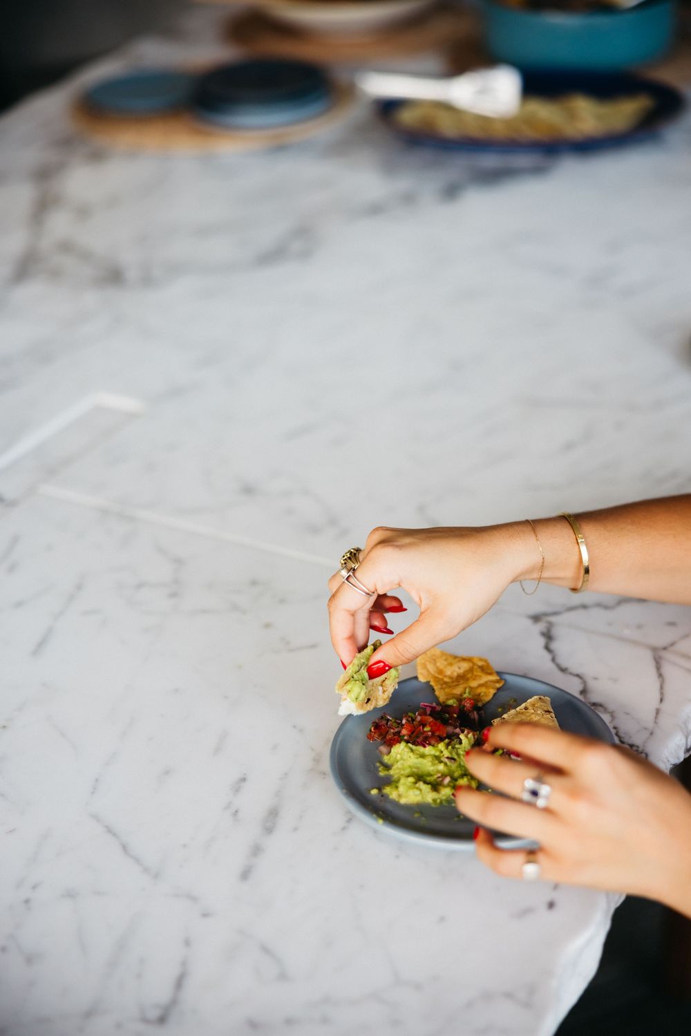 Hands with jewelry arranging ingredients on a burger over a gray marble countertop.