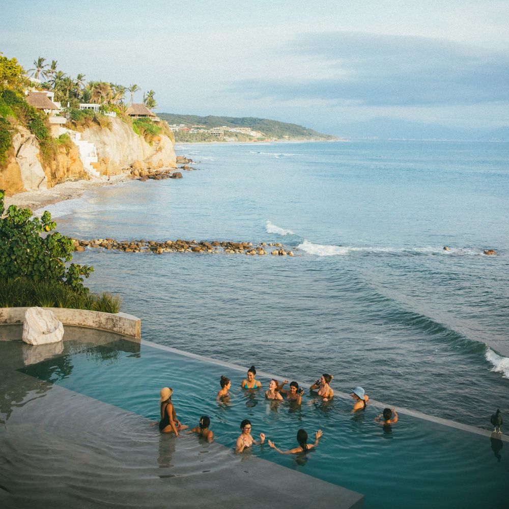 Group of friends enjoying a scenic swim in a tranquil ocean cove with rocky cliffs and blue waters.