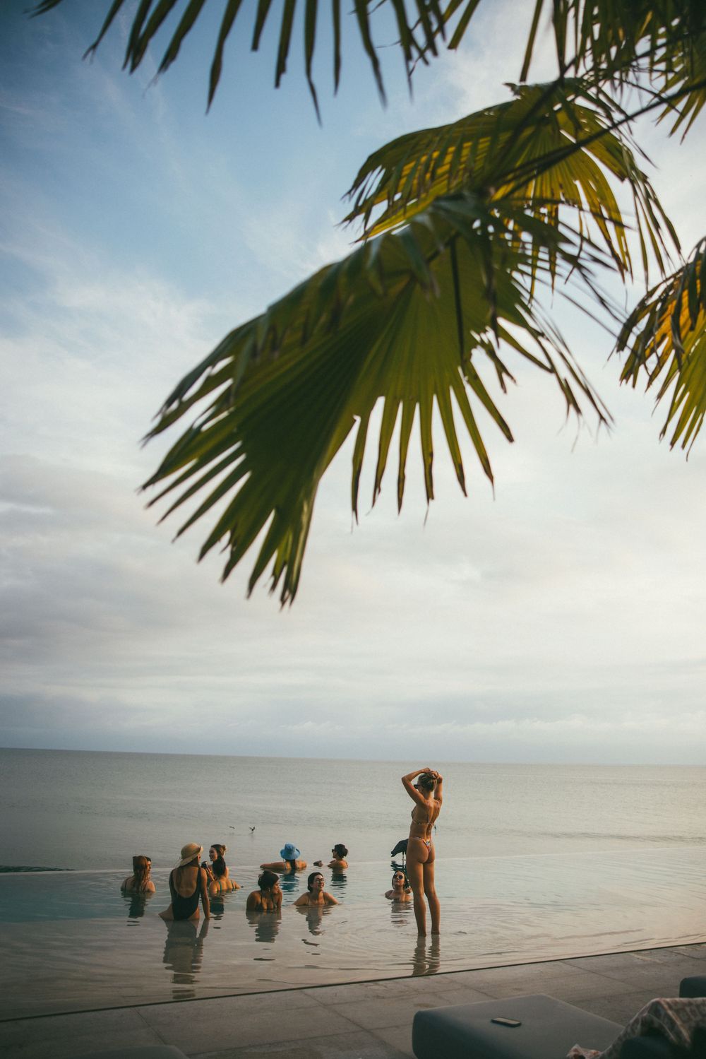 People enjoying a tropical beach scene with palm fronds overhead and calm ocean waters stretching to the horizon.