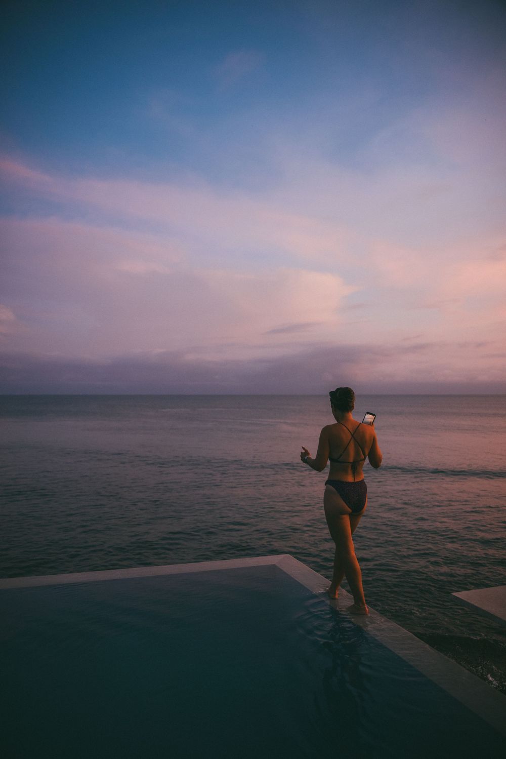 A silhouetted figure stands on a beach pier at sunset overlooking a tranquil ocean with pink and purple sky.
