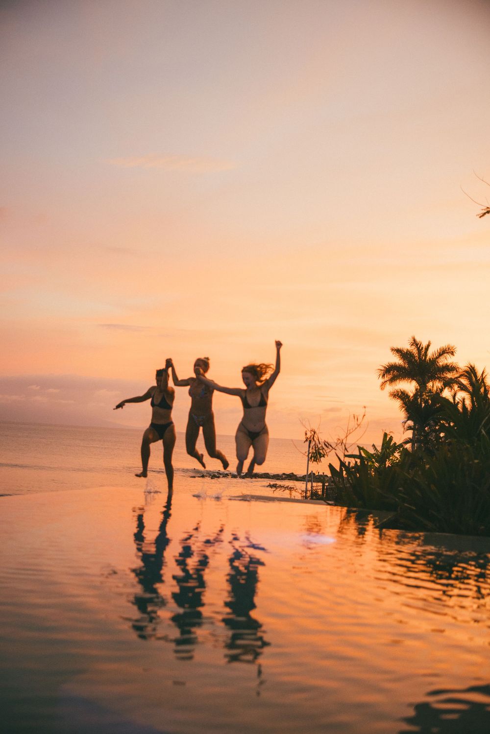 Silhouettes of people jumping and playing in a tropical infinity pool during a vibrant orange sunset.