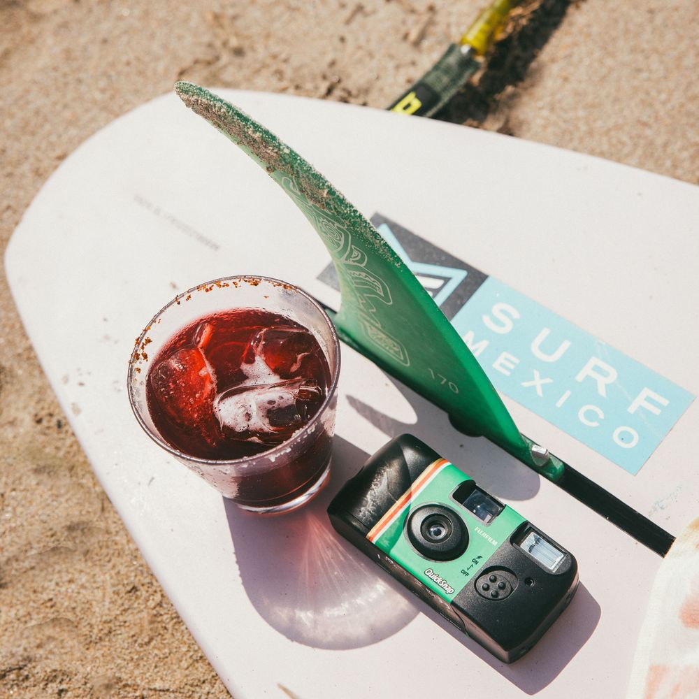 Bottle of Lubi sparkling water and glass with ice on sandy beach surface in bright sunlight.