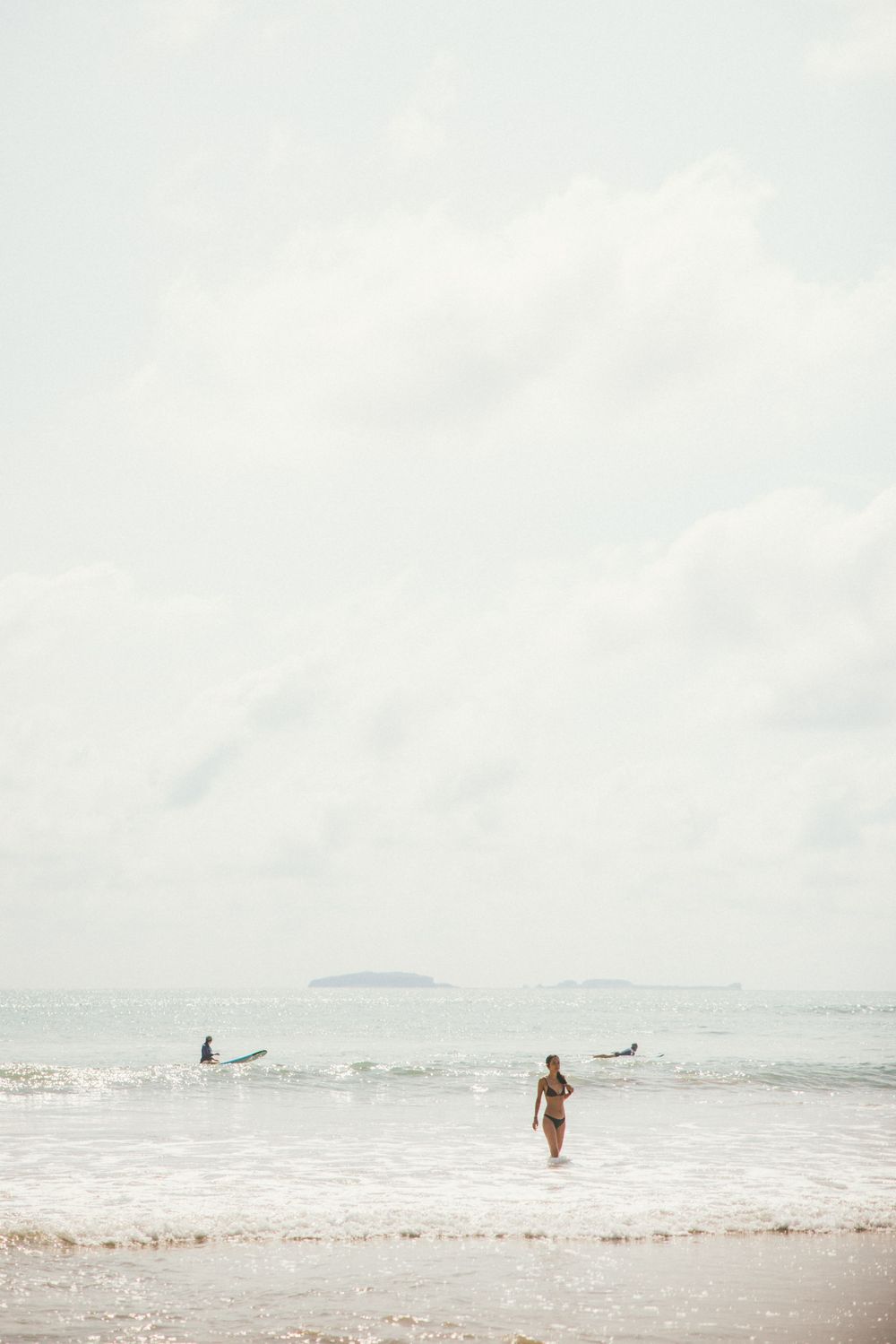 Surfers walk along a misty beach shoreline with rolling waves under an overcast sky.