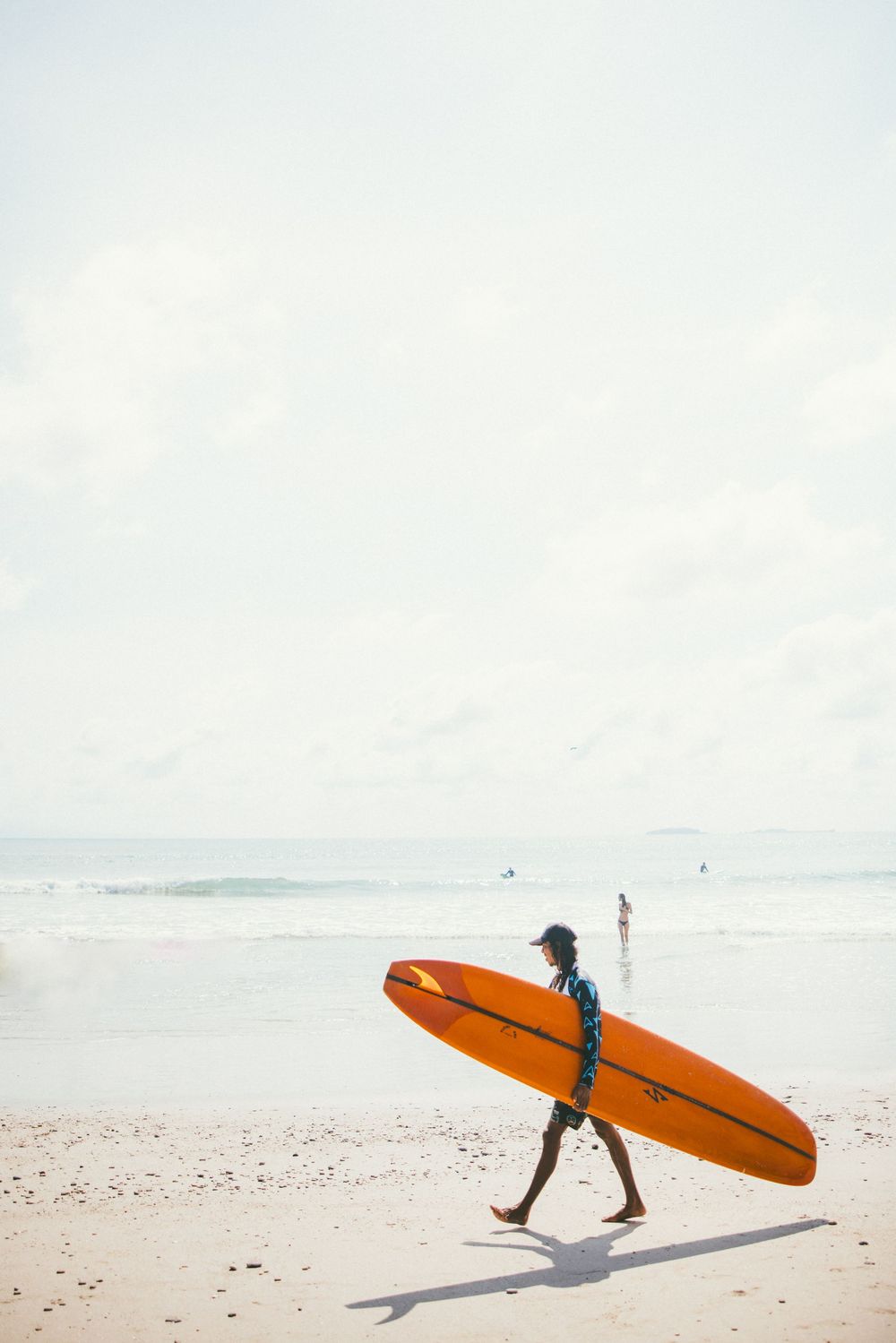 Person walking on a sandy beach carrying an orange surfboard against a bright sky.