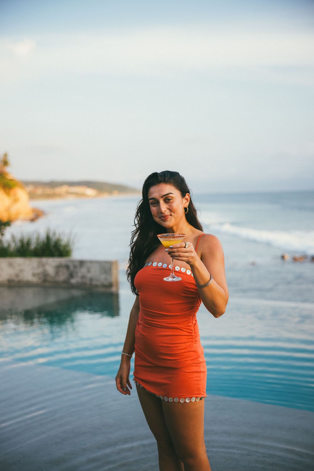 A sequence of joyful moments captured of someone in an orange swimsuit posing by an oceanfront infinity pool.