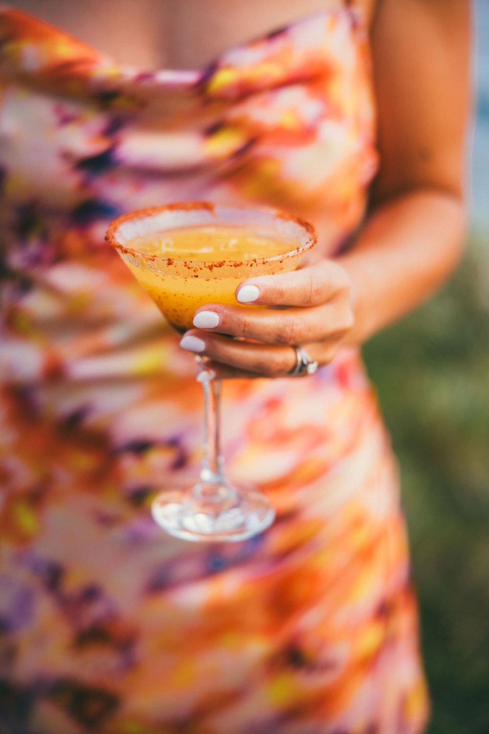 A colorful margarita glass with sugar-rimmed edge is held against a blurred orange and pink floral background.