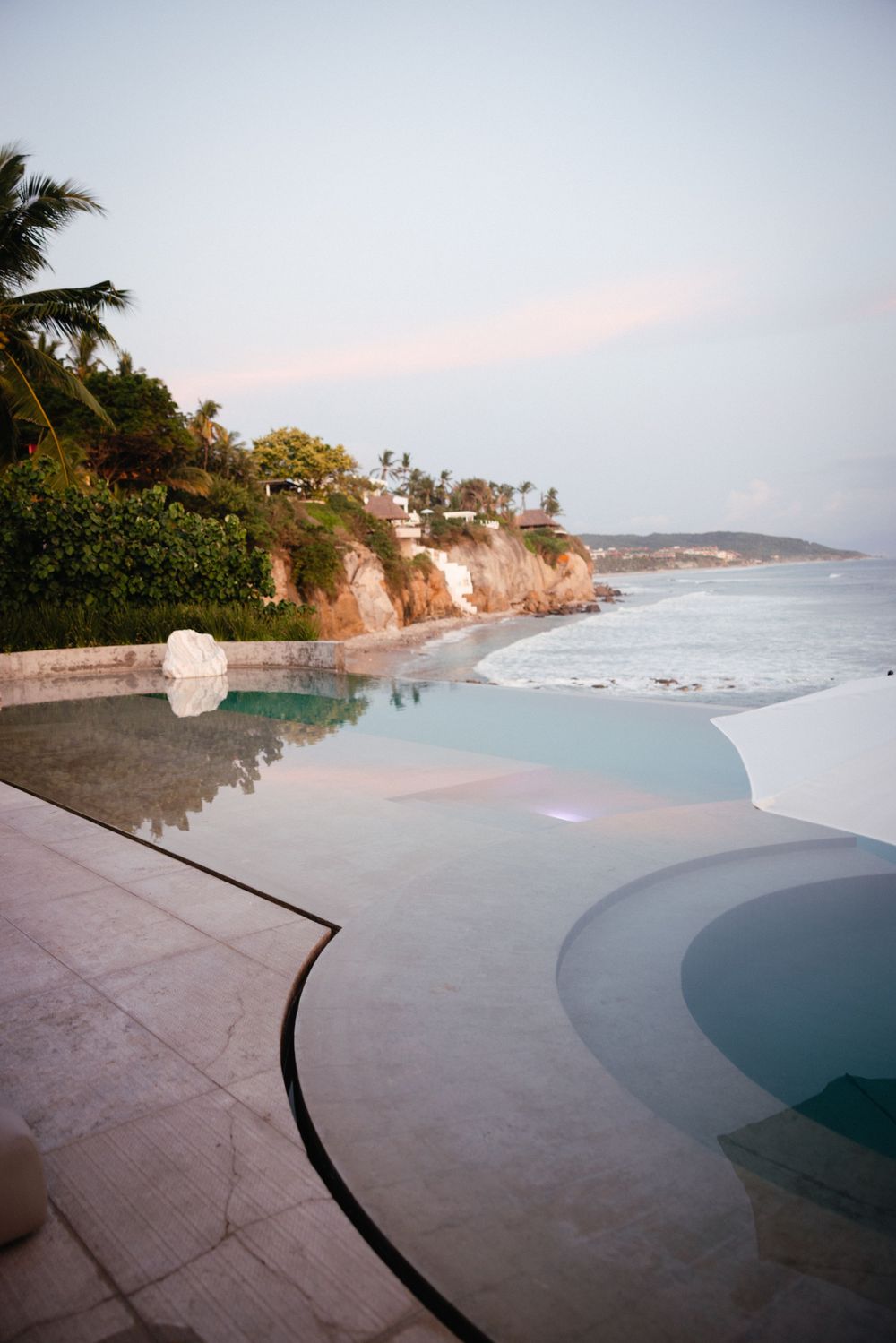 An infinity pool overlooks a dramatic oceanfront cliff at sunset with palm trees and rocky coastline in view.