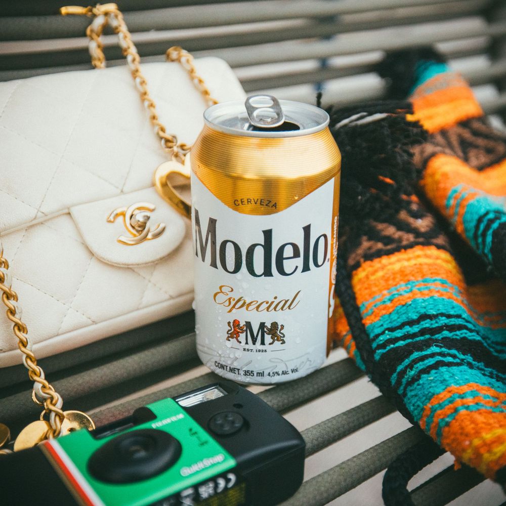 A row of beer cans including Modelo and other brands arranged on a wooden surface with orange and teal decorative elements.