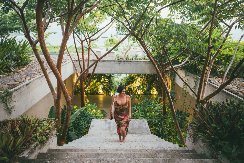 A person in a pink dress walks down a scenic stone staircase surrounded by lush tropical greenery.