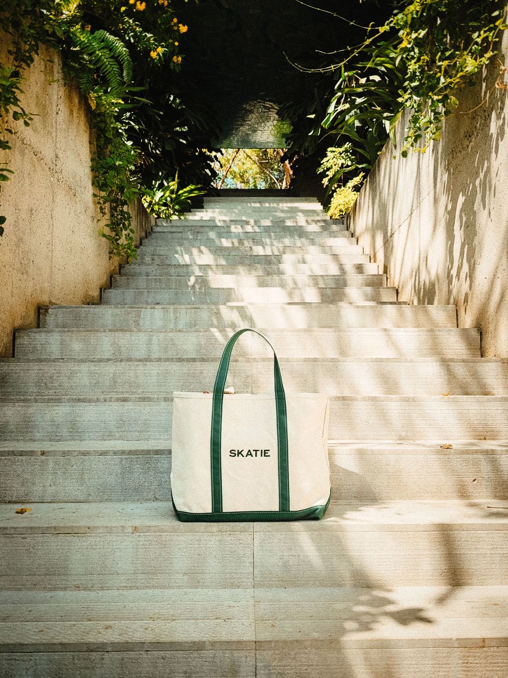 White canvas tote bag sitting on concrete steps with dappled sunlight and greenery overhead.