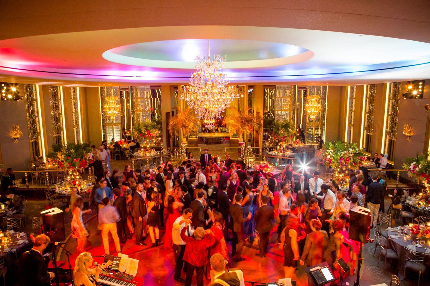 Colorful lights illuminate a crowded dance floor at an elegant ballroom reception venue with chandeliers and decorative walls.