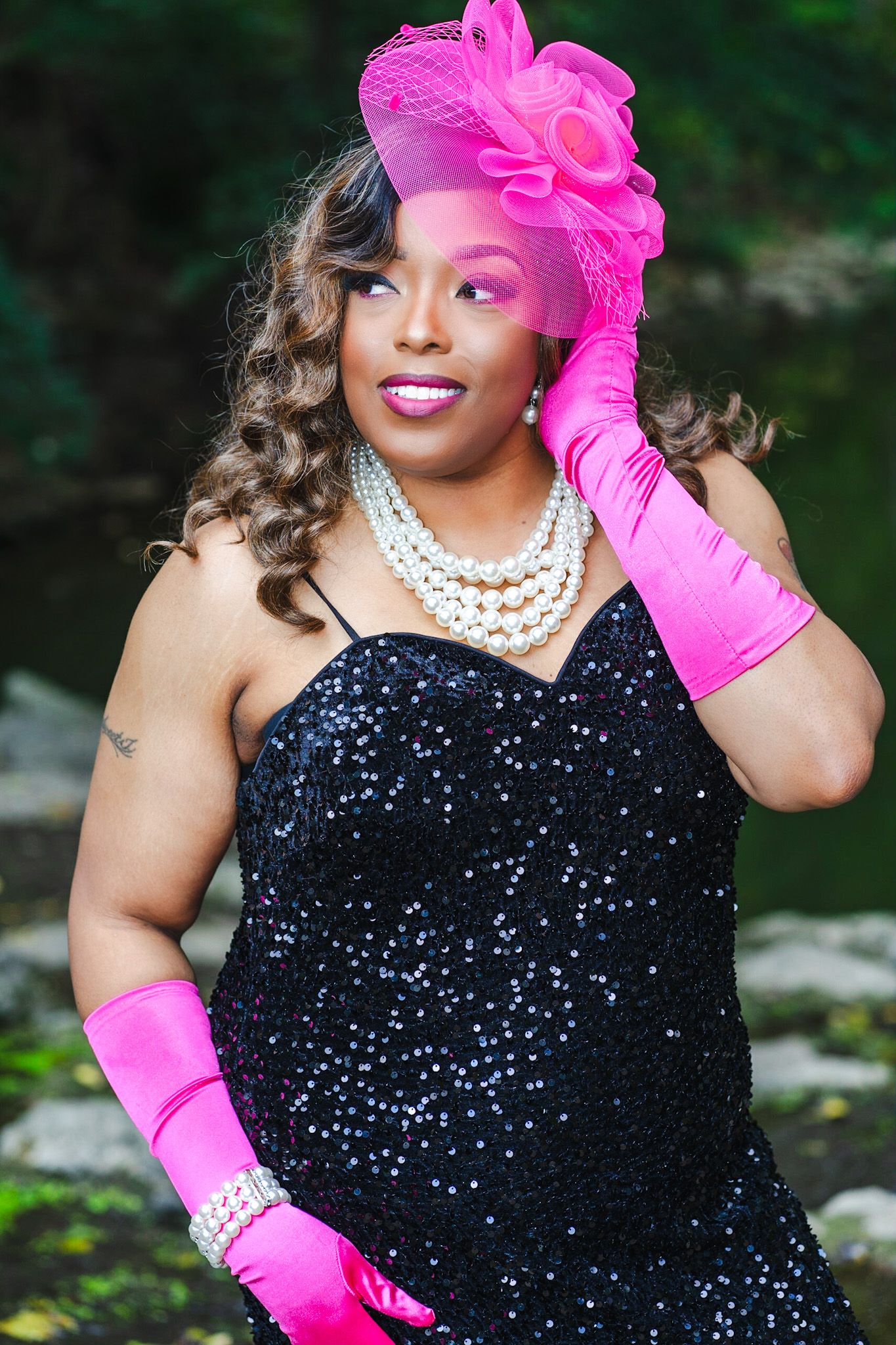 Elegant evening look with sparkling black dress, pink gloves and layered pearl necklace in garden setting.