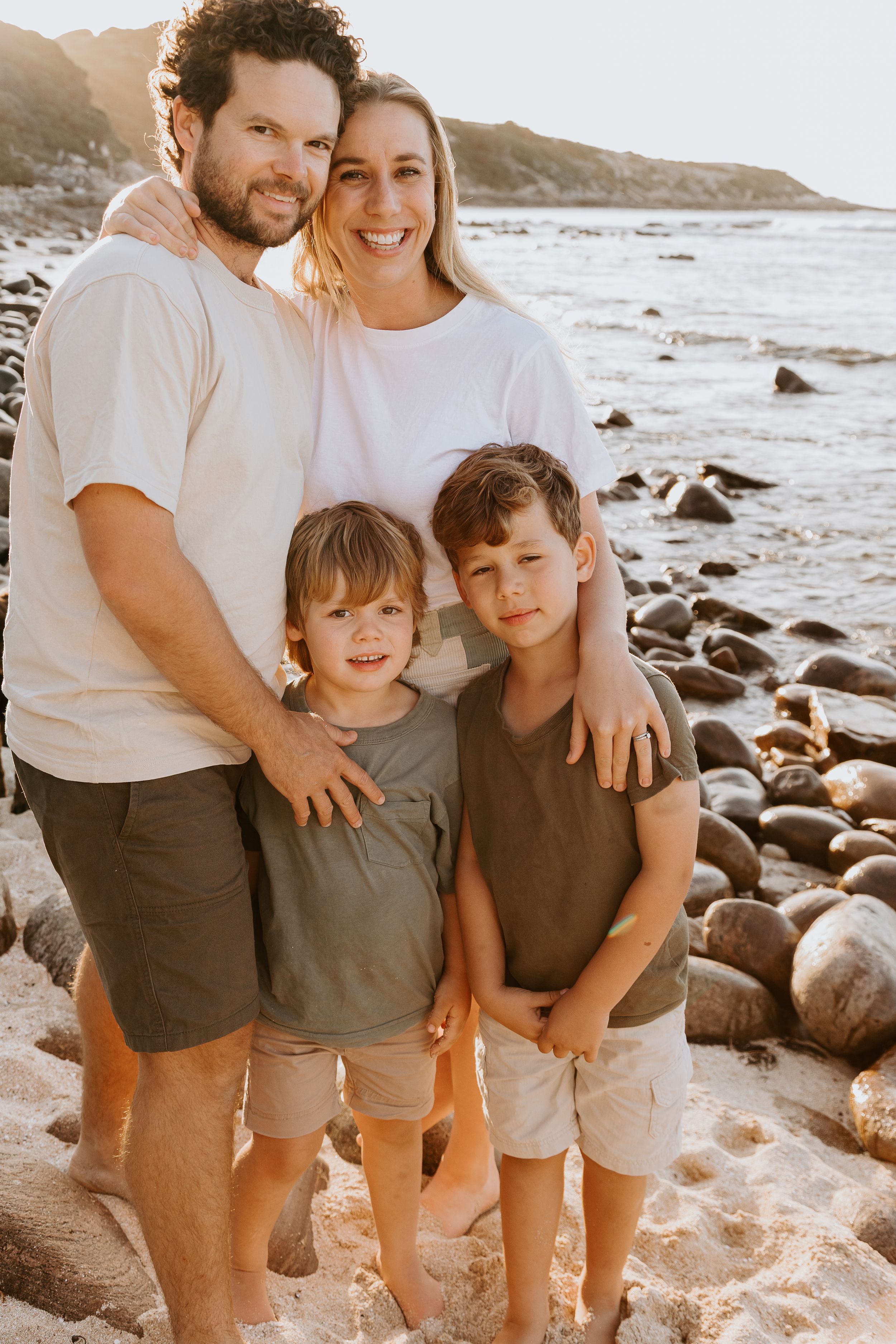 A family wearing coordinated white and gray outfits poses together on a rocky beach at sunset.