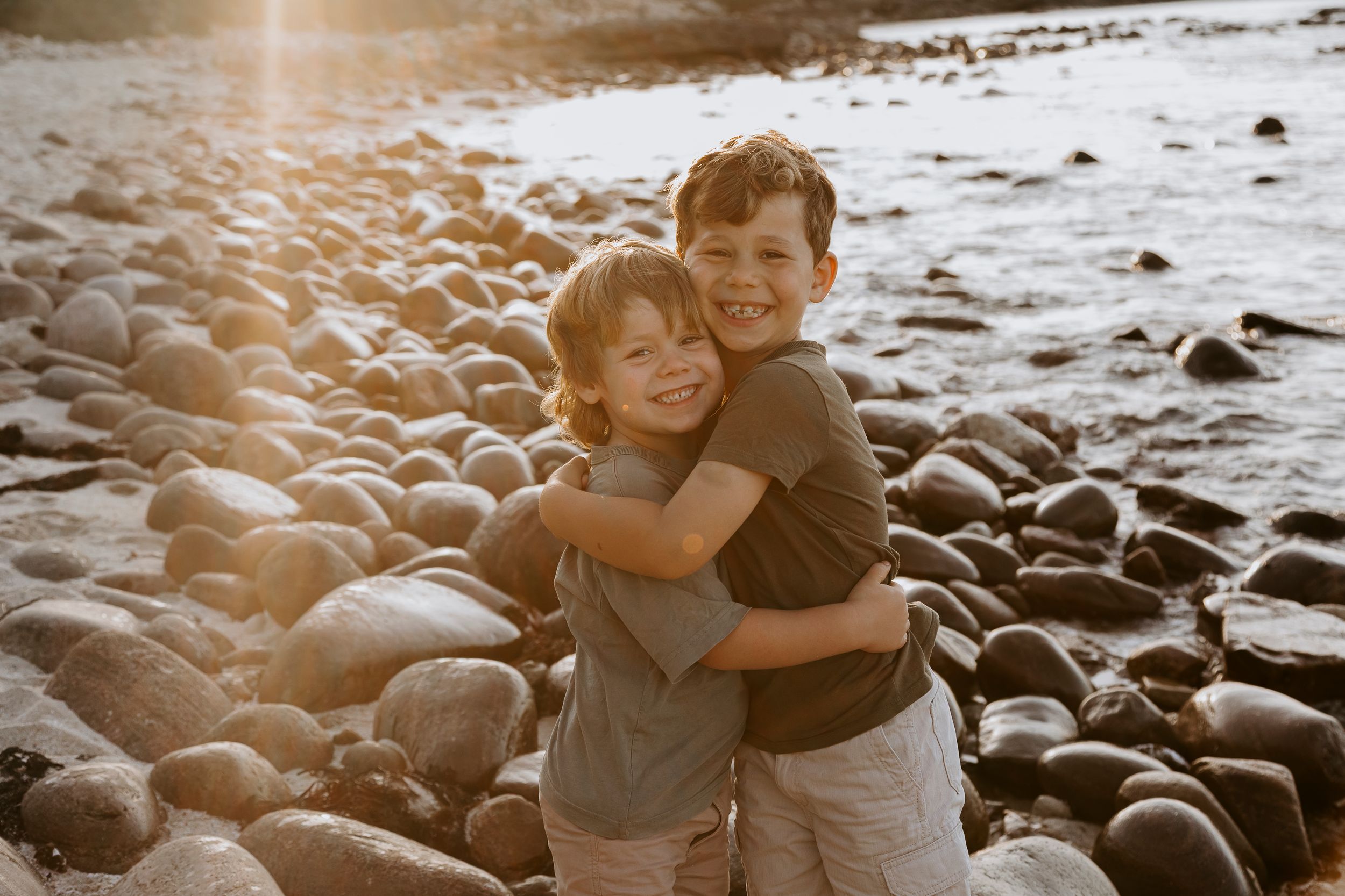 A couple shares an intimate moment on a rocky beach at sunset with warm golden light filtering through the scene.