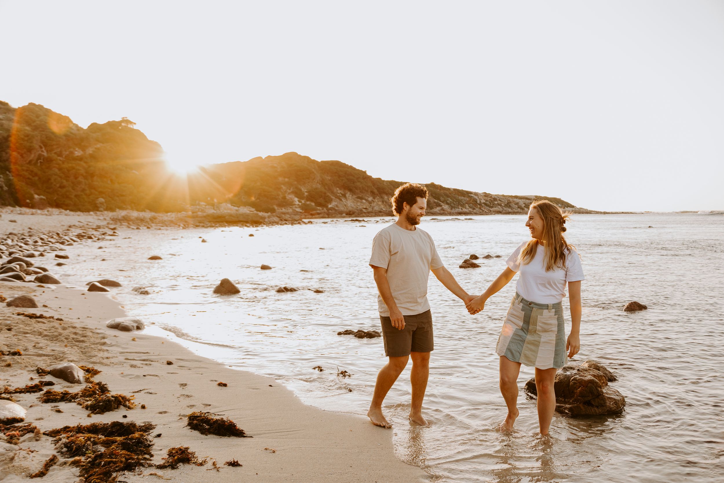 Couple walking hand in hand along a scenic beach at sunset with waves crashing on rocky shore.