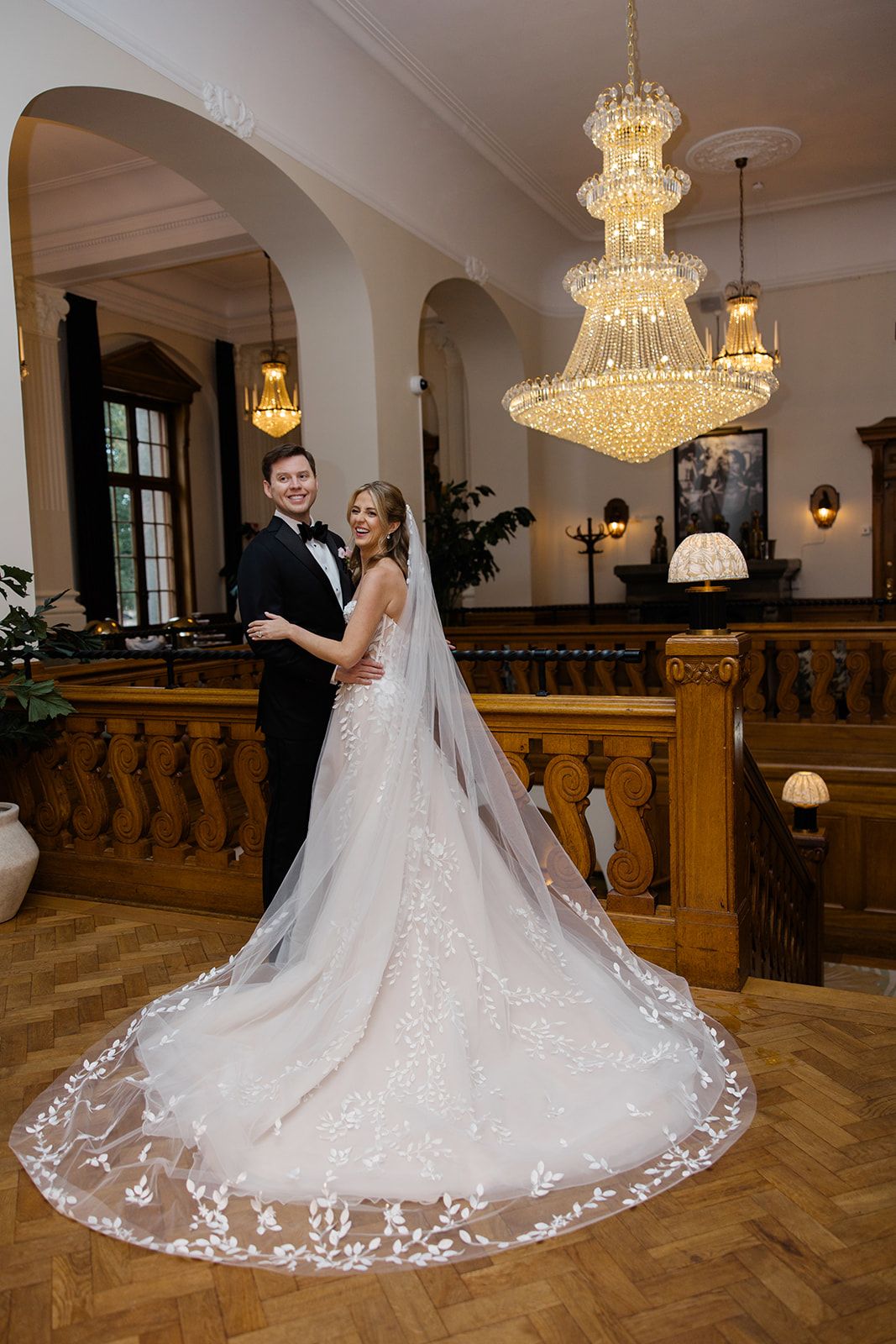 Elegant wedding couple poses beneath ornate crystal chandelier on hardwood floor with long lace veil trailing behind.