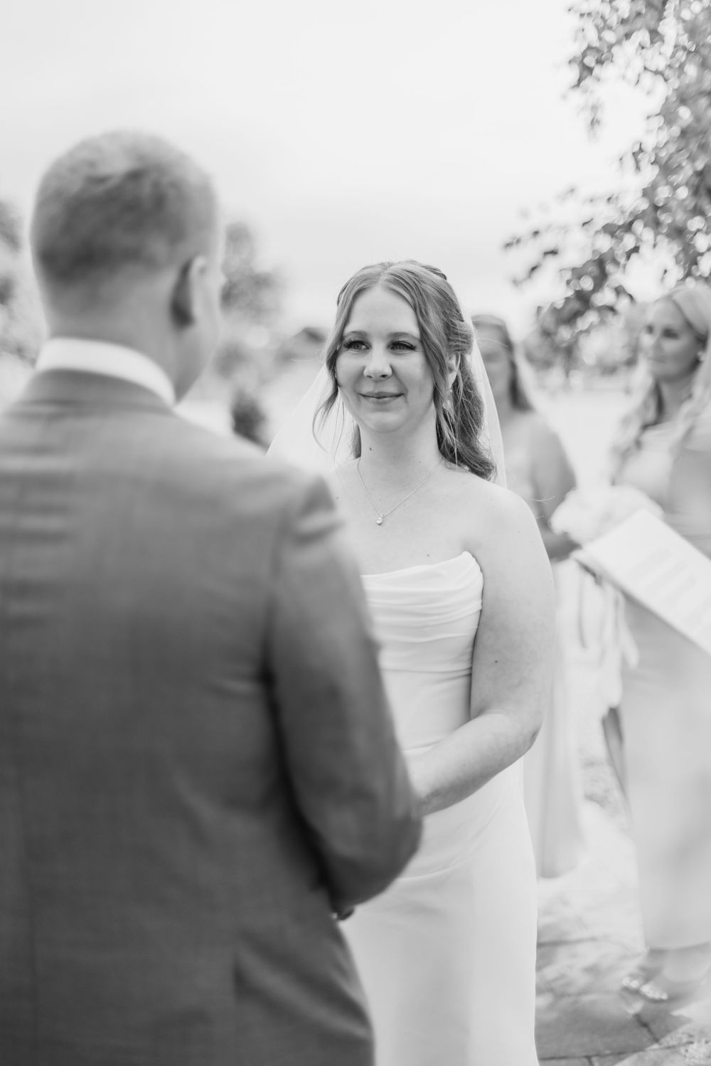 A black and white wedding ceremony photo showing a couple exchanging vows outdoors.