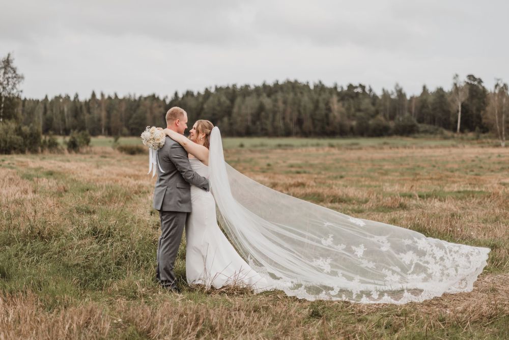 A couple in wedding attire walks through a scenic meadow with mountains and forest in the background.