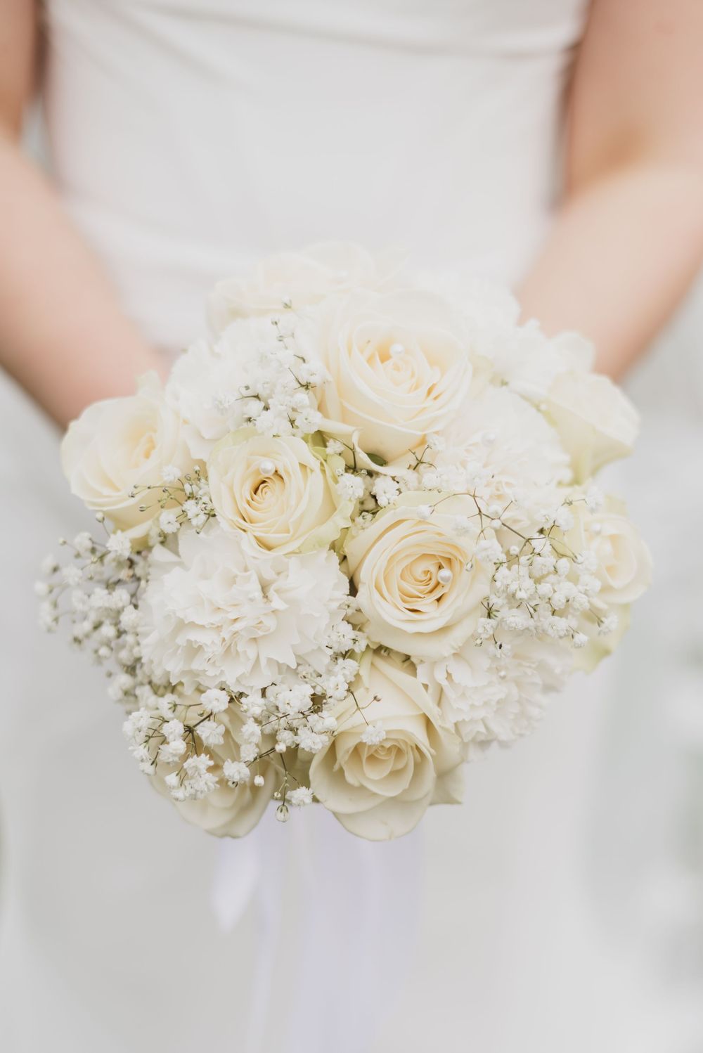 Elegant white wedding bouquet with cream roses and baby's breath flowers against a light background.