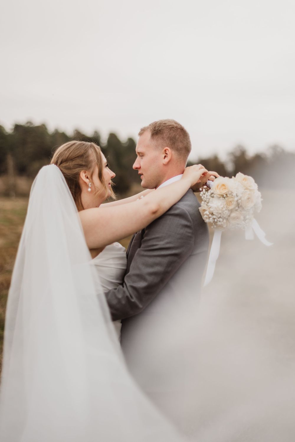 A romantic wedding couple embraces outdoors in a misty field while the bride holds a white bouquet.