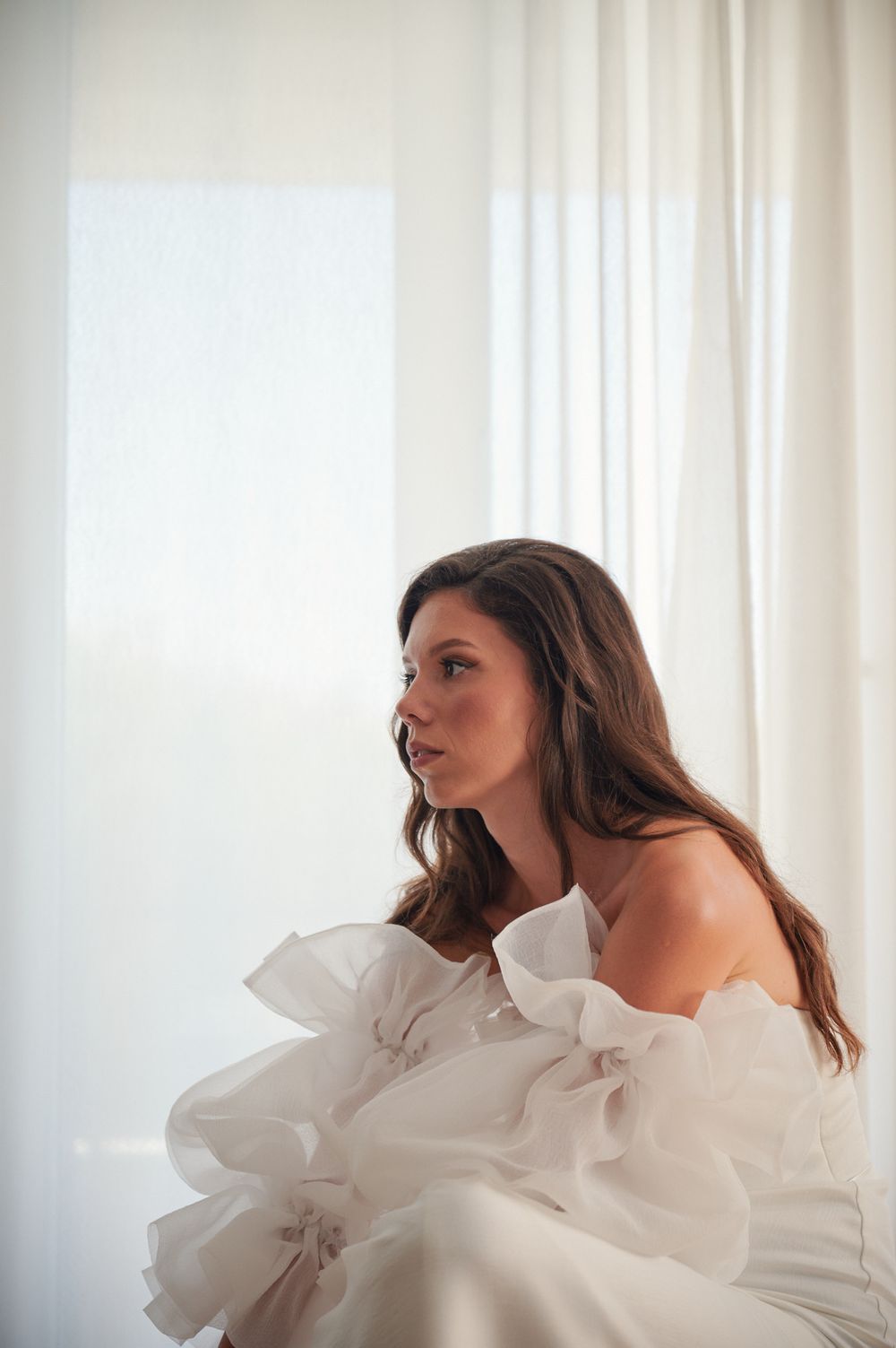 Woman in white ruffled off-shoulder dress poses elegantly against light curtains.