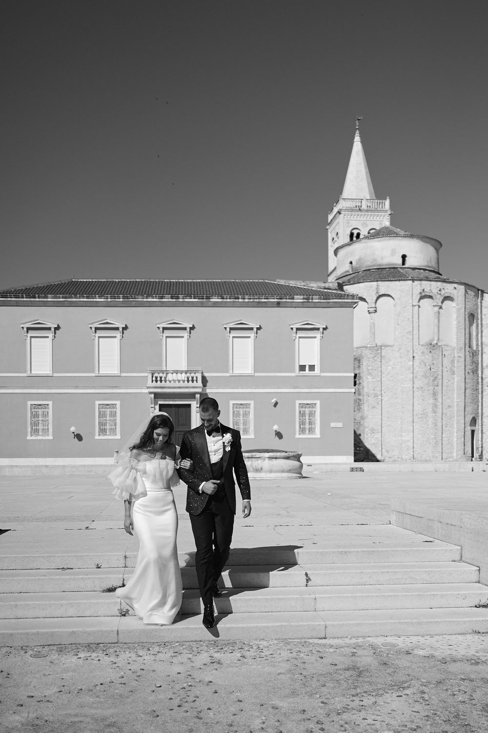 Black and white photo of couple walking along historic stone building with architectural details.