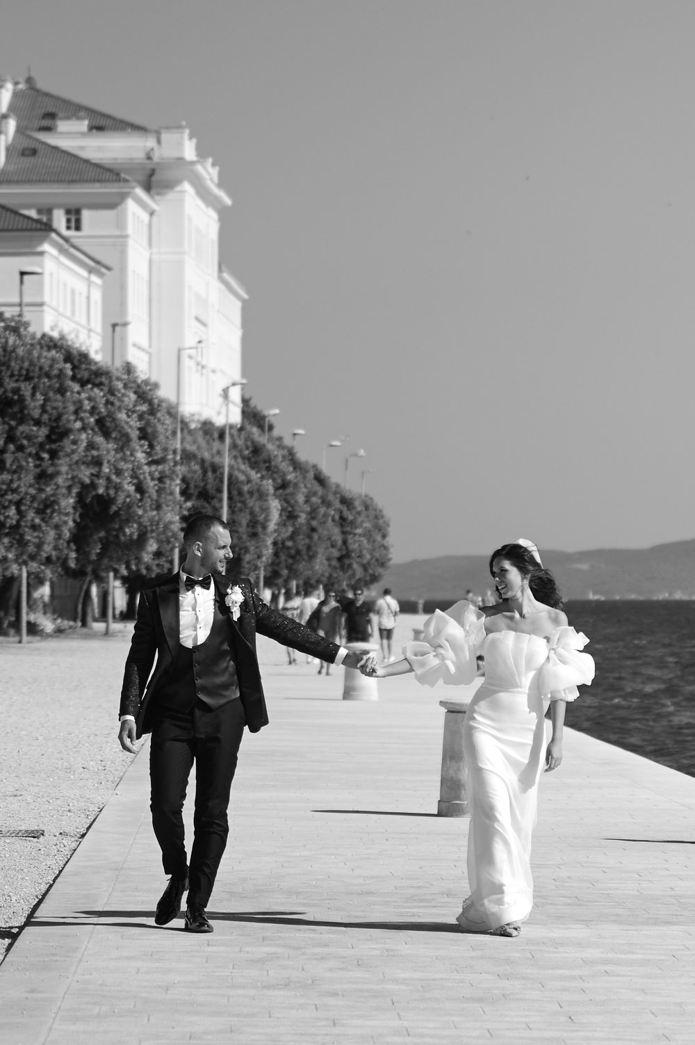 A couple in wedding attire walks along a seaside promenade on a sunny day.