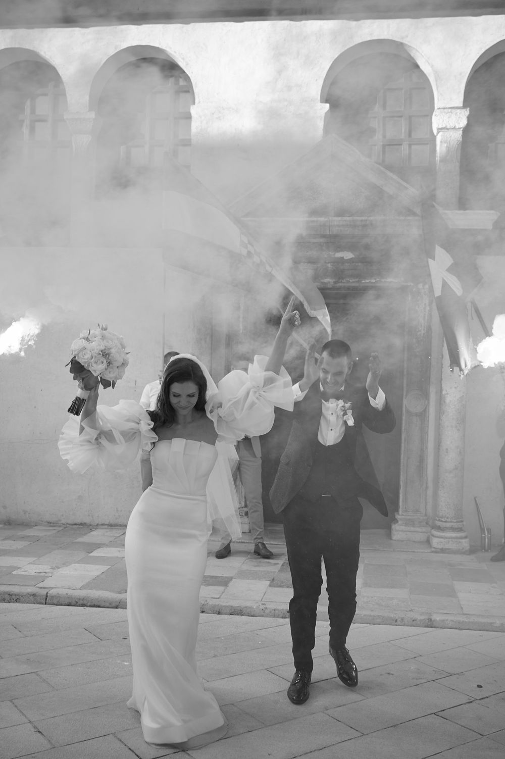 A wedding party walks through atmospheric smoke in front of a historic stone building.