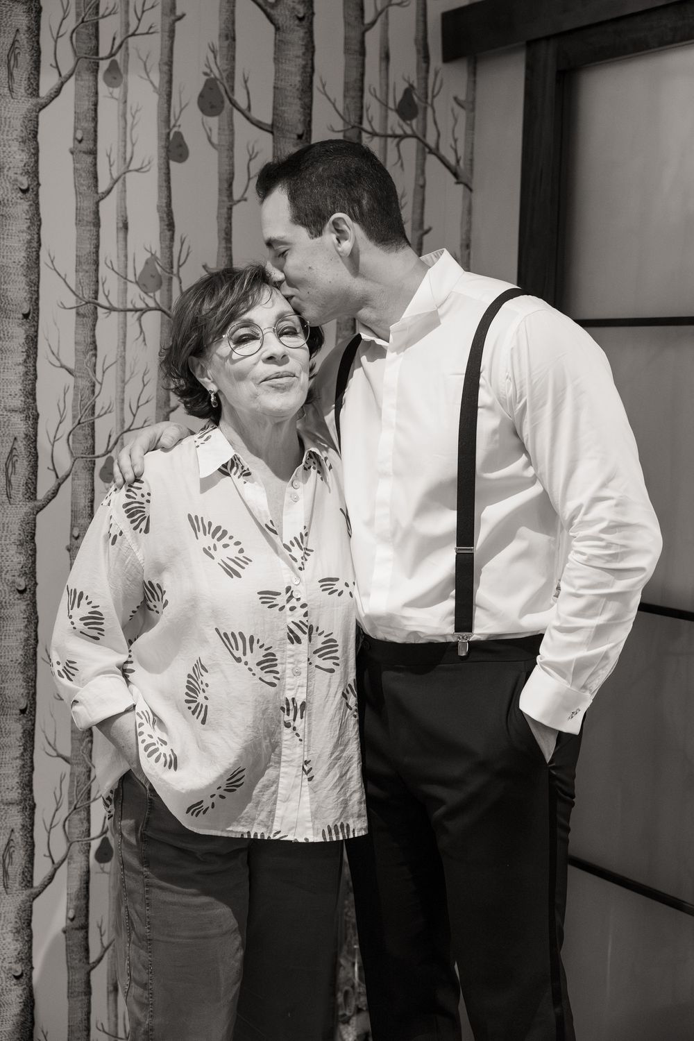 A black and white photo of two people sharing an intimate moment against a wallpapered wall with birch tree pattern.