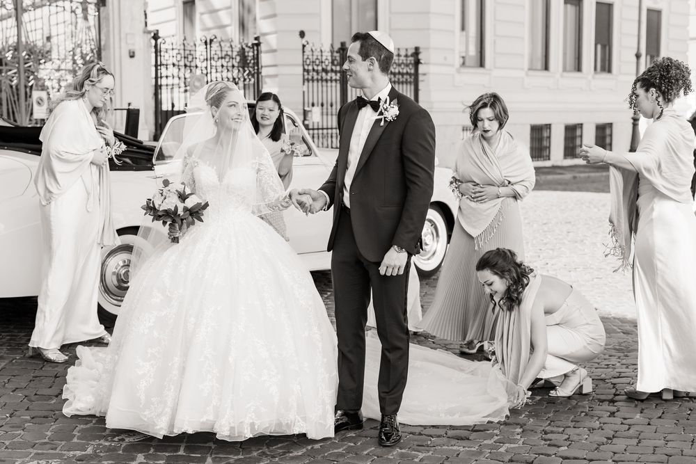 A vintage black and white wedding scene shows a bride in a full skirt gown with attendants on a cobblestone street.