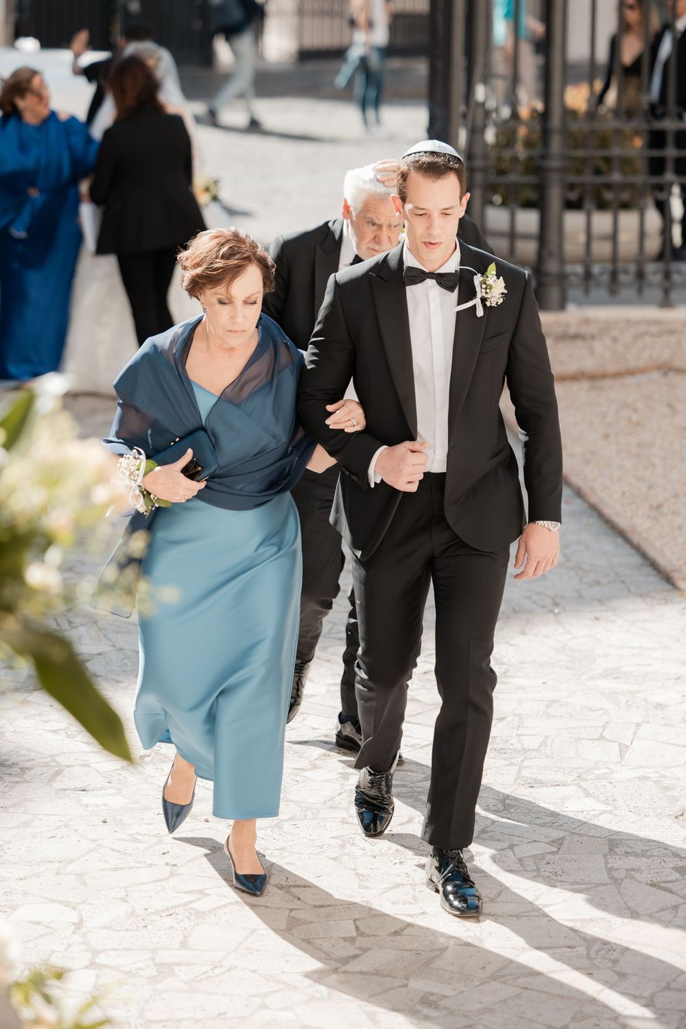 A groom in a black tuxedo walks arm in arm with a woman in a blue dress at an elegant outdoor wedding ceremony.