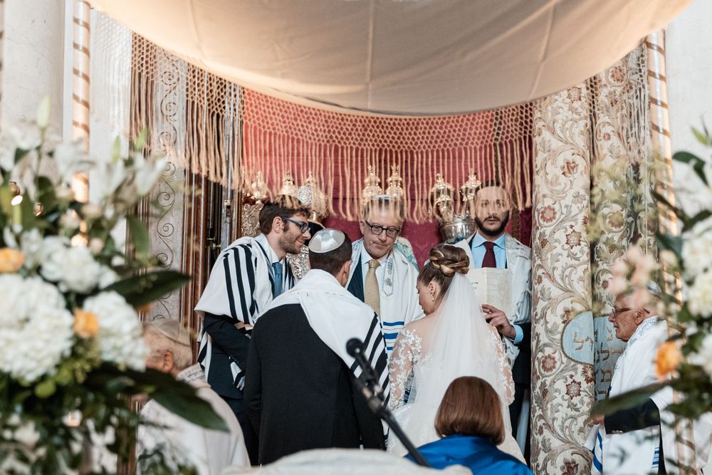 Jewish wedding ceremony takes place under a traditional chuppah decorated with white fabric and floral arrangements.