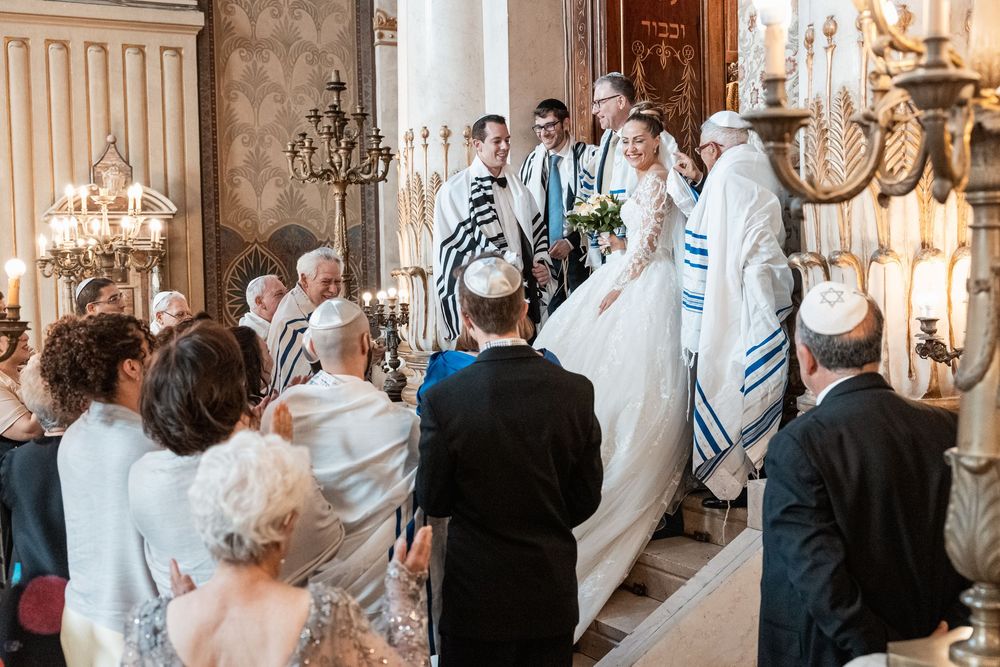 A Jewish wedding ceremony takes place under elegant chandeliers with attendees wearing traditional prayer shawls.