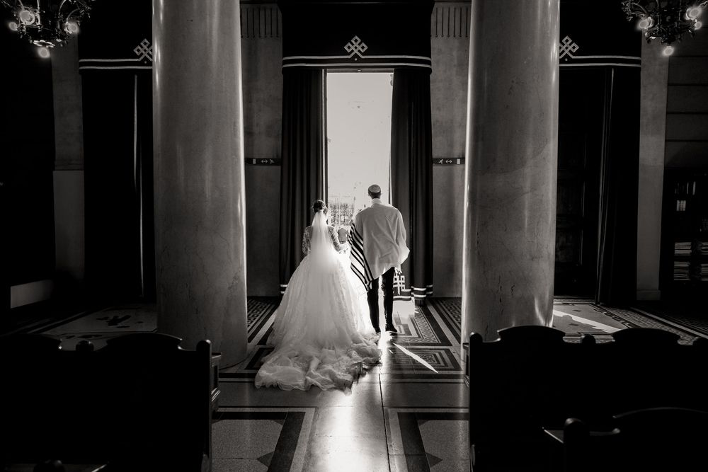 A bride in a white wedding gown stands elegantly between classical columns in a dramatic black and white photograph.