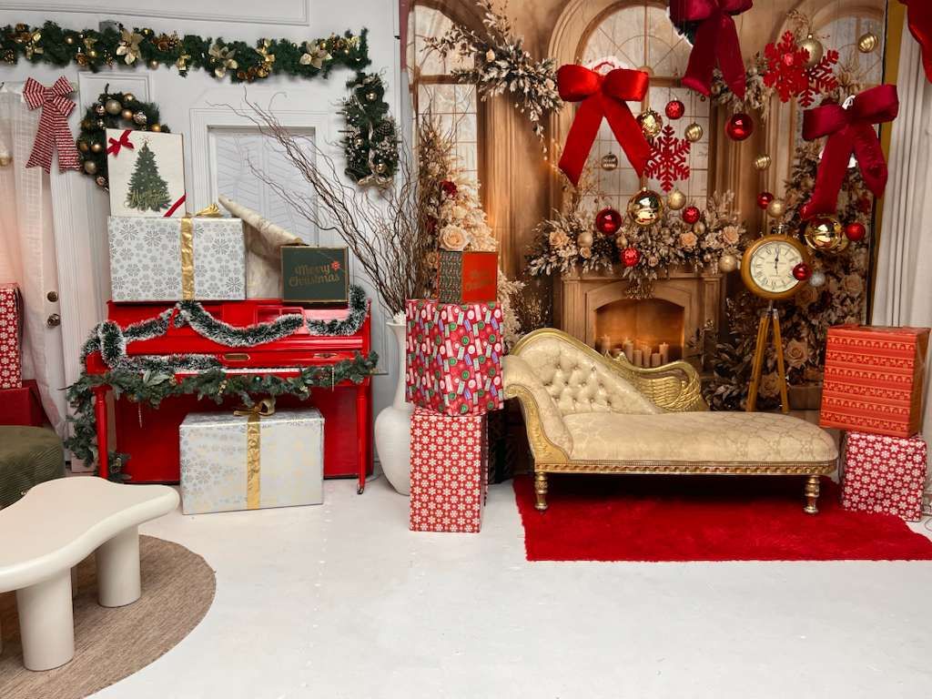 Festive Christmas photo studio set with red and gold decorations, wrapped gifts, chaise lounge, and decorated fireplace.