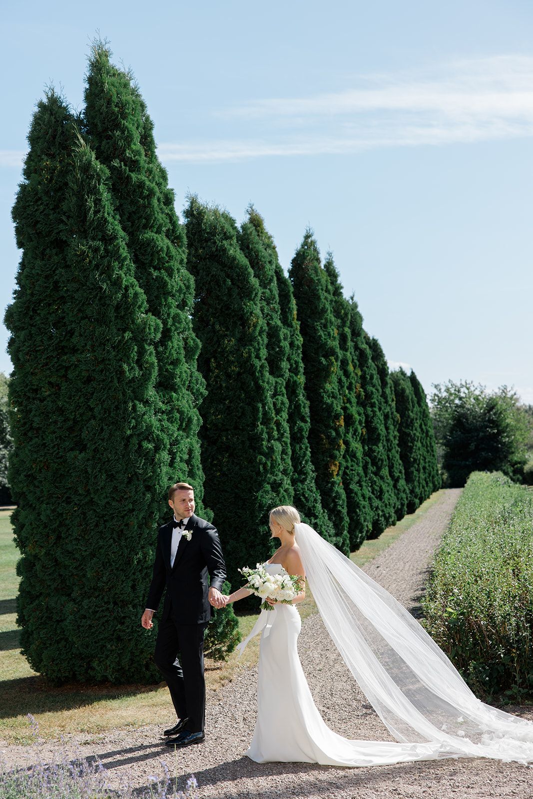 A romantic wedding couple shares an intimate moment among cypress trees lining a garden path.