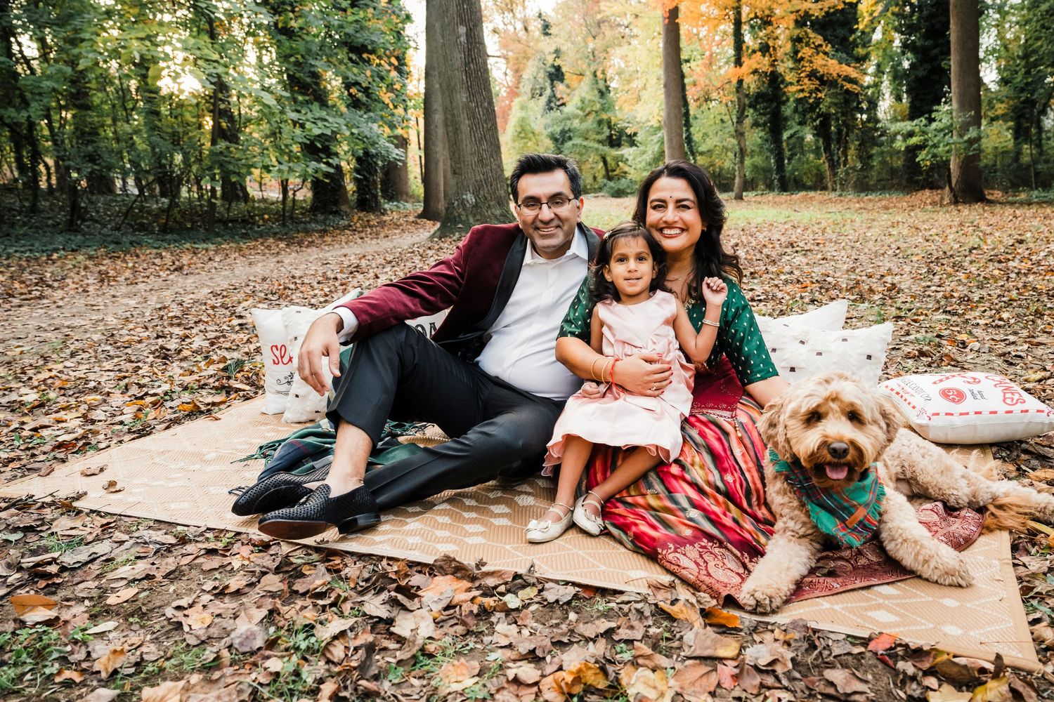 A couple and their dog pose together on fallen autumn leaves in a wooded setting.