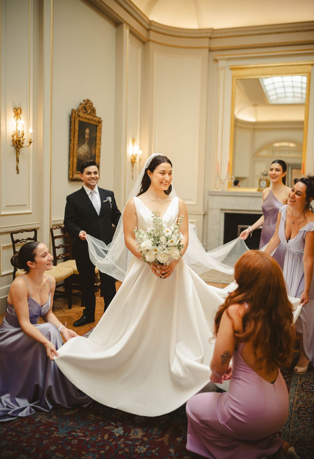 Bridesmaids in purple dresses helping arrange the bride's wedding gown.
