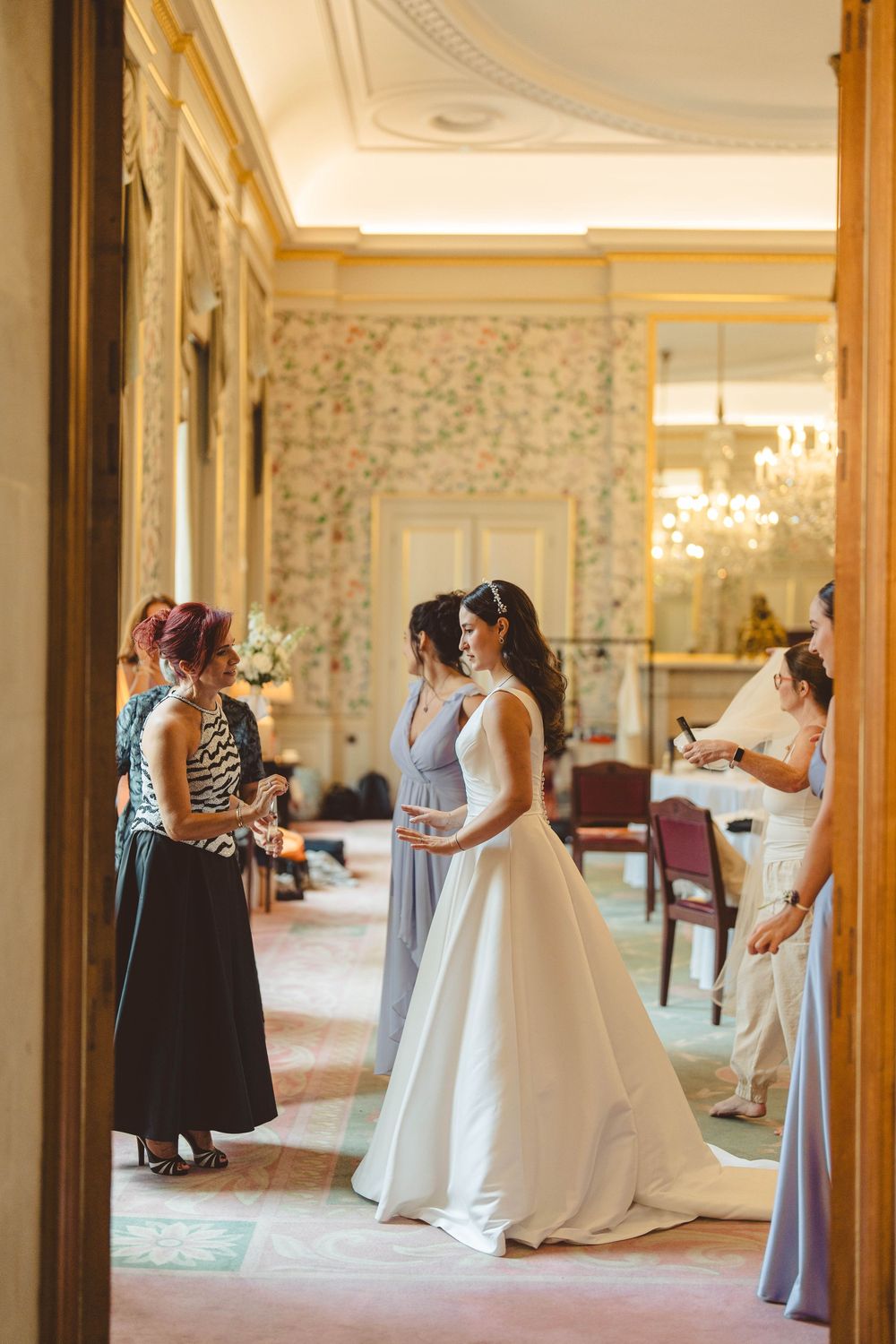 Wedding party getting ready in elegant room with floral wallpaper and chandeliers.