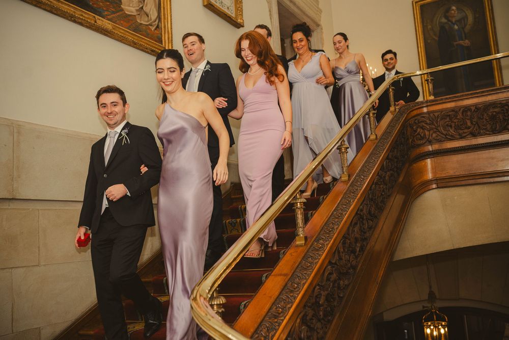 Wedding party members in formal attire descending ornate wooden staircase.
